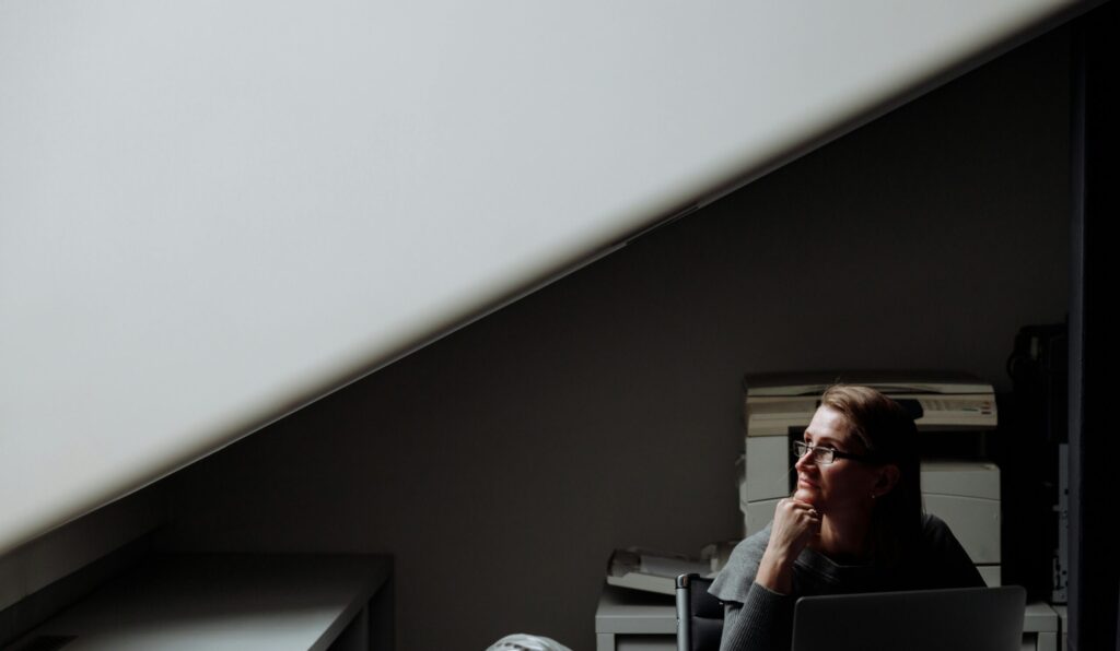 A woman sits at a desk in a dimly lit office, contemplating while using a laptop.