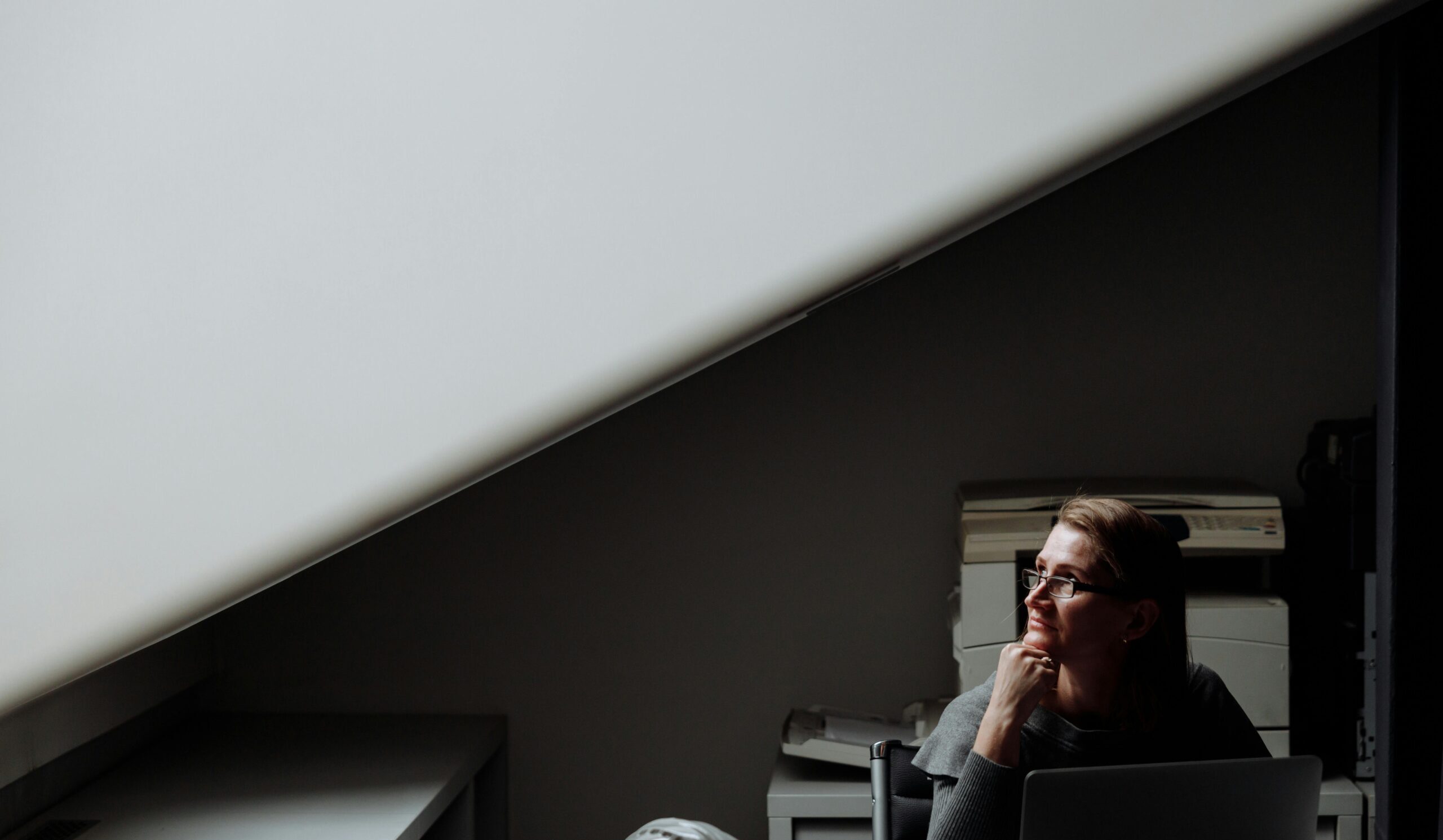 A woman sits at a desk in a dimly lit office, contemplating while using a laptop.
