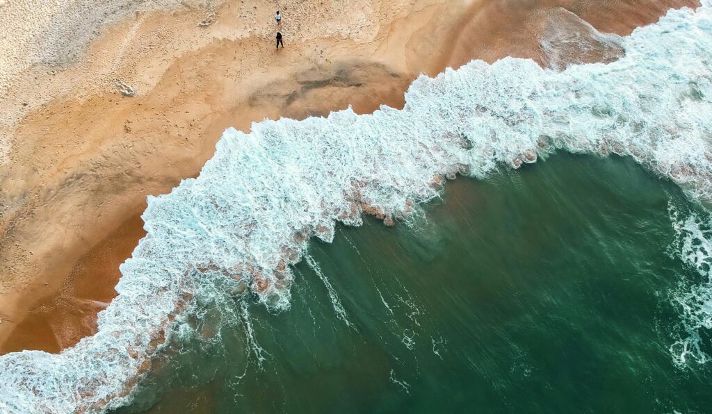 Aerial shot of ocean waves crashing onto a sandy beach with two people in the distance.