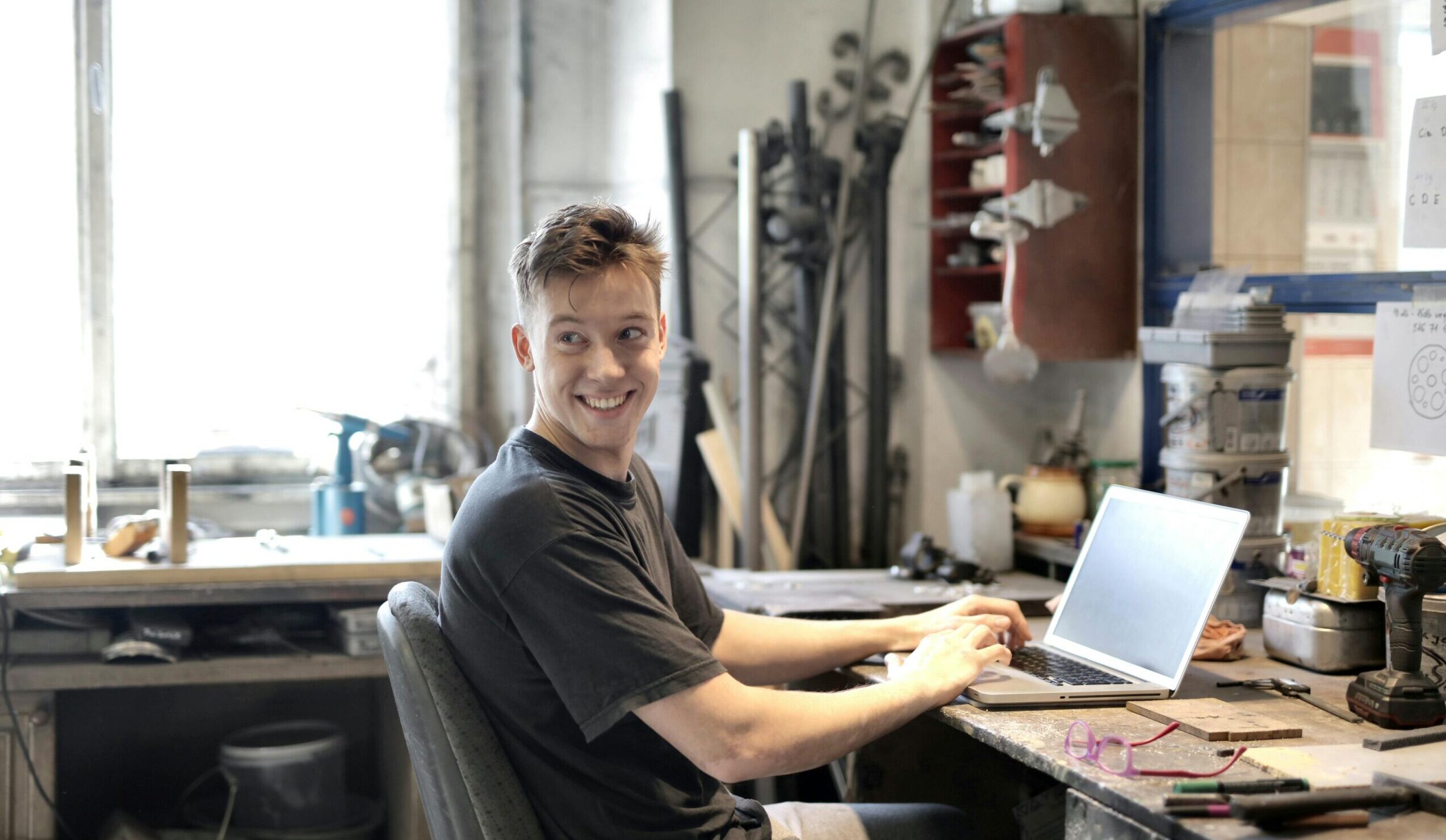 Young man smiling while using laptop in a creative workspace with tools around.